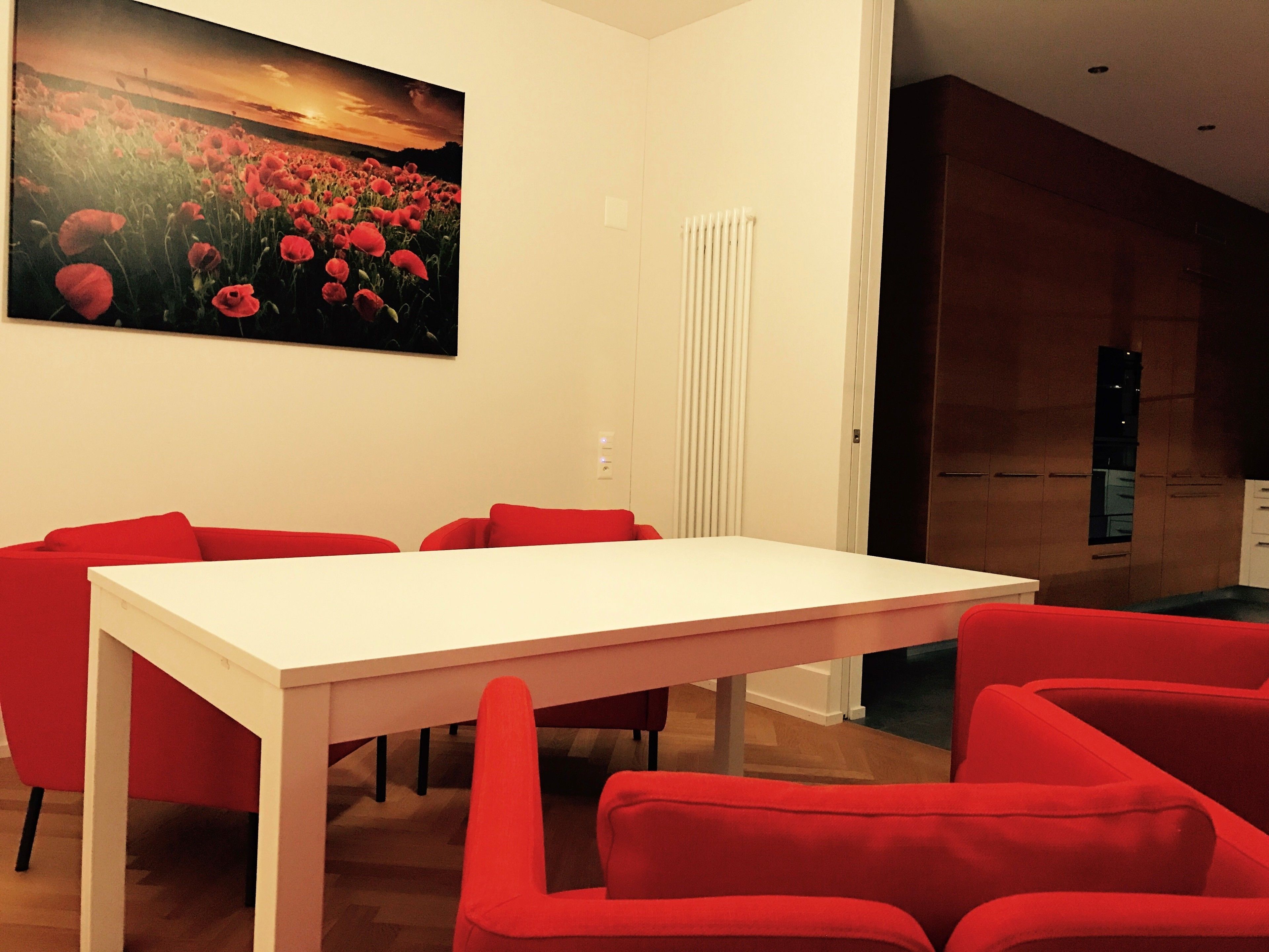 A dining area with a white table and red chairs, a large painting of a field of red flowers on the wall, and a modern kitchen in the background.