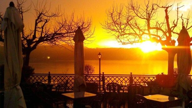 An outdoor seating area with tables and chairs, a statue, and a view of the sunset over a body of water. - Villa Degli Angeli (Castel Gandolfo)