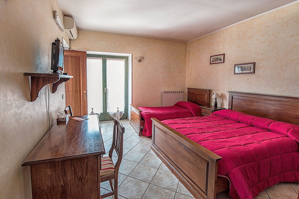 A bedroom with a wooden desk, two beds with red bedding, a wooden headboard, a bedside table with a lamp, and a wall-mounted air conditioner.