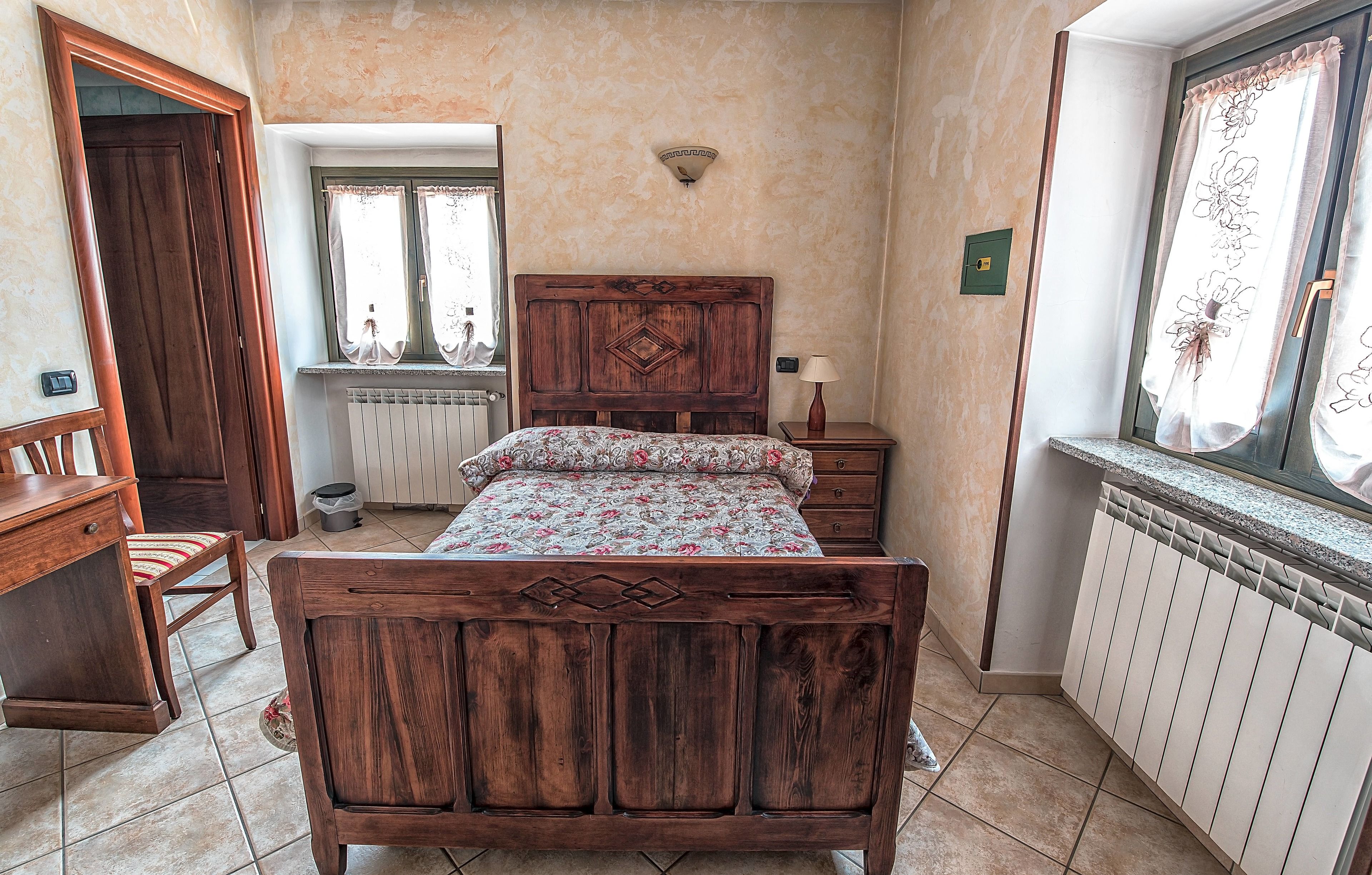 A bedroom with a wooden bed, a nightstand, a chair, a window with curtains, and a radiator.