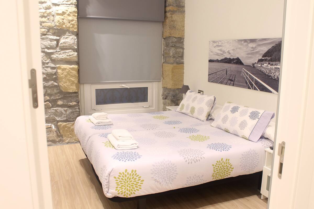 A bedroom with a stone wall, a bed with a floral bedspread, a window with a blind, and a framed picture on the wall.
