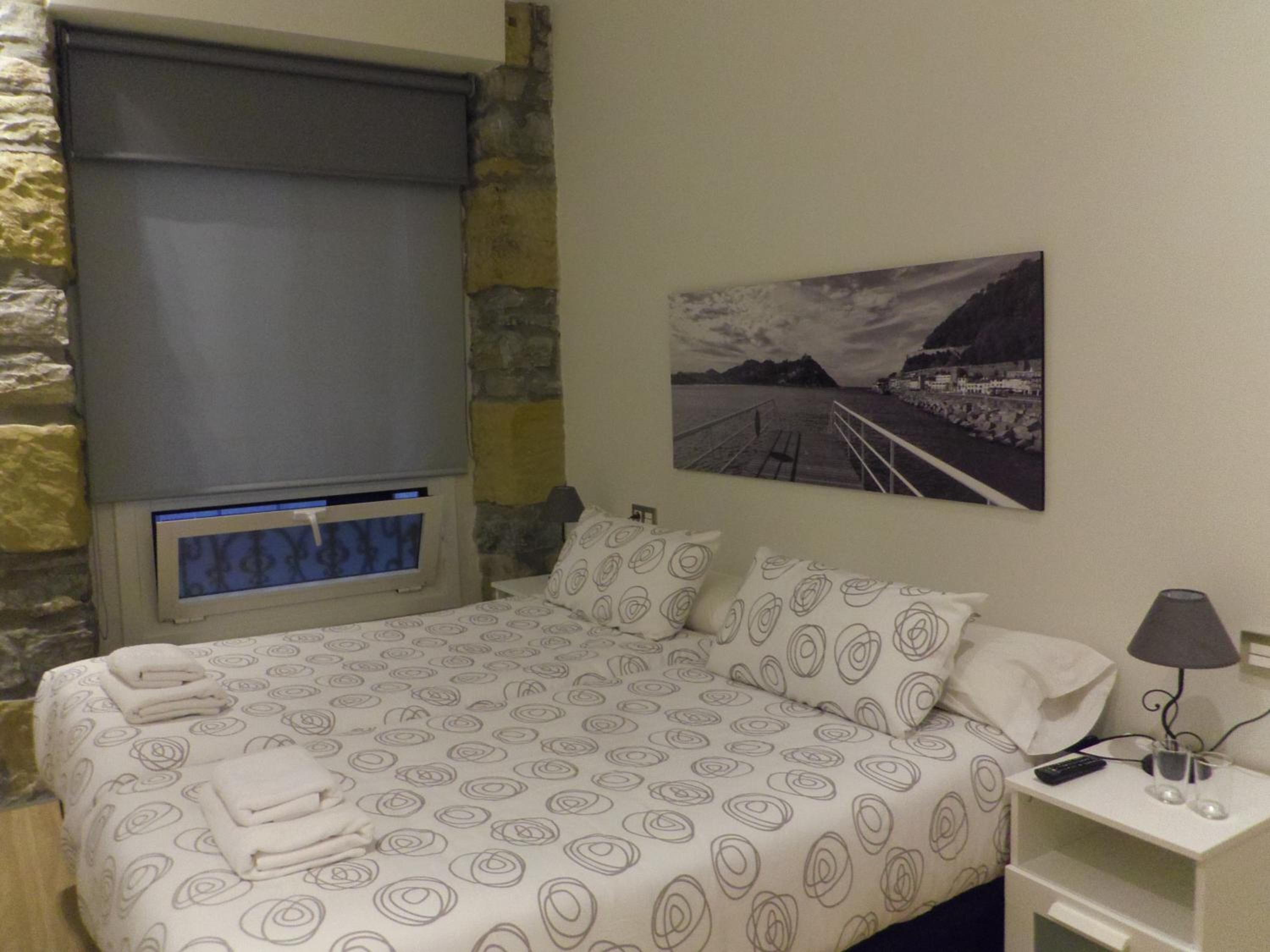 Un dormitorio con cama, ventana con vistas, un cuadro en la pared y mesita de noche con lámpara.