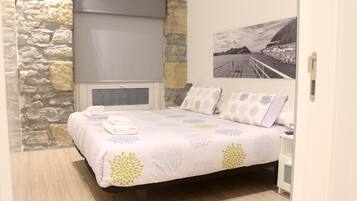 Une chambre avec un mur en pierre, un lit recouvert d’une couvre-lit à motifs floraux, une fenêtre avec un store et un tableau représentant un paysage côtier.