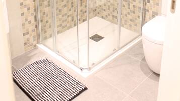Un box de ducha con paredes de mosaico cerámico, un inodoro blanco y un felpudo negro en la entrada.