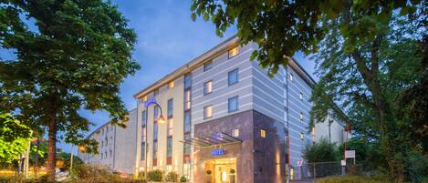 A modern hotel with a glass entrance, surrounded by greenery and a cobblestone path.