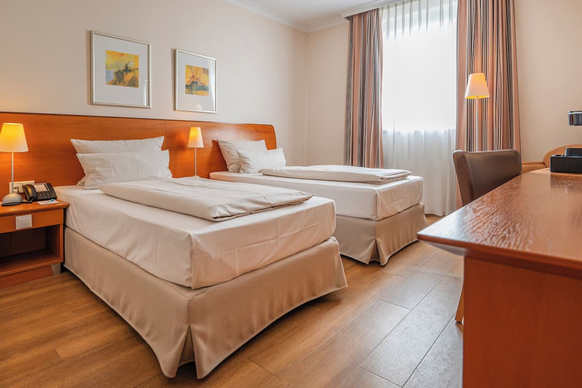 A hotel room with two beds, a wooden desk, and a chair. There are two bedside lamps, a telephone, and artwork on the wall.