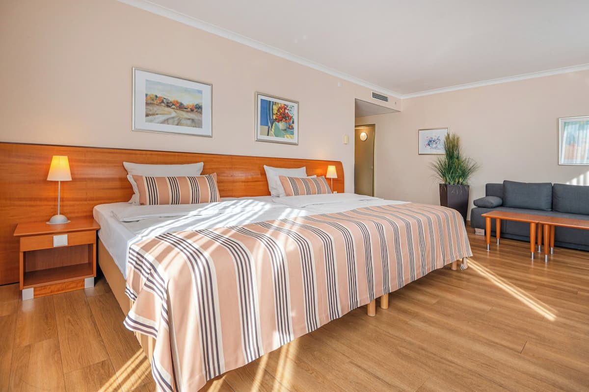 A hotel room with a large bed, a wooden nightstand, a bedside table with a lamp, a grey sofa, a wooden coffee table, and a potted plant.