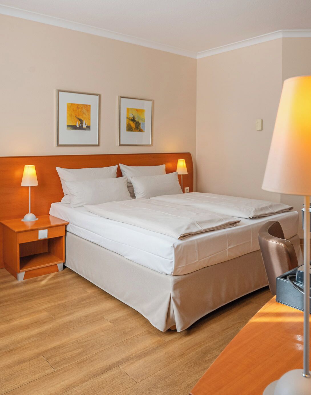 A hotel room with a large bed, two bedside tables with lamps, a wooden floor, and two framed pictures on the wall.