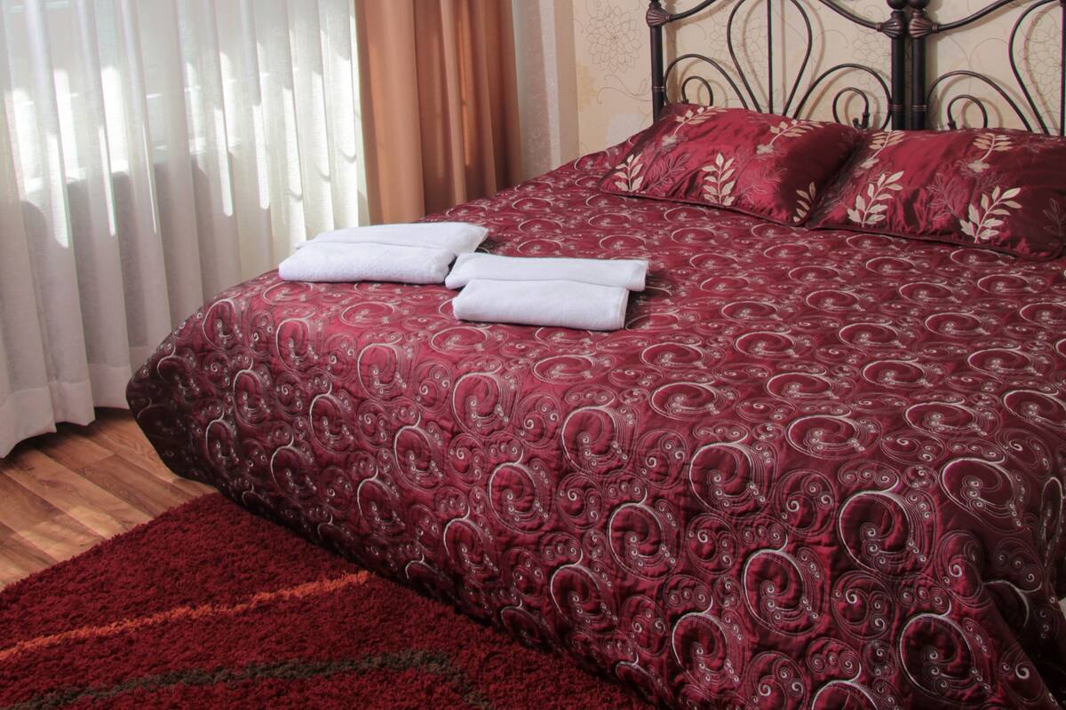 A neatly made bed with a patterned bedspread and pillows, a red carpet, and a window with sheer curtains.