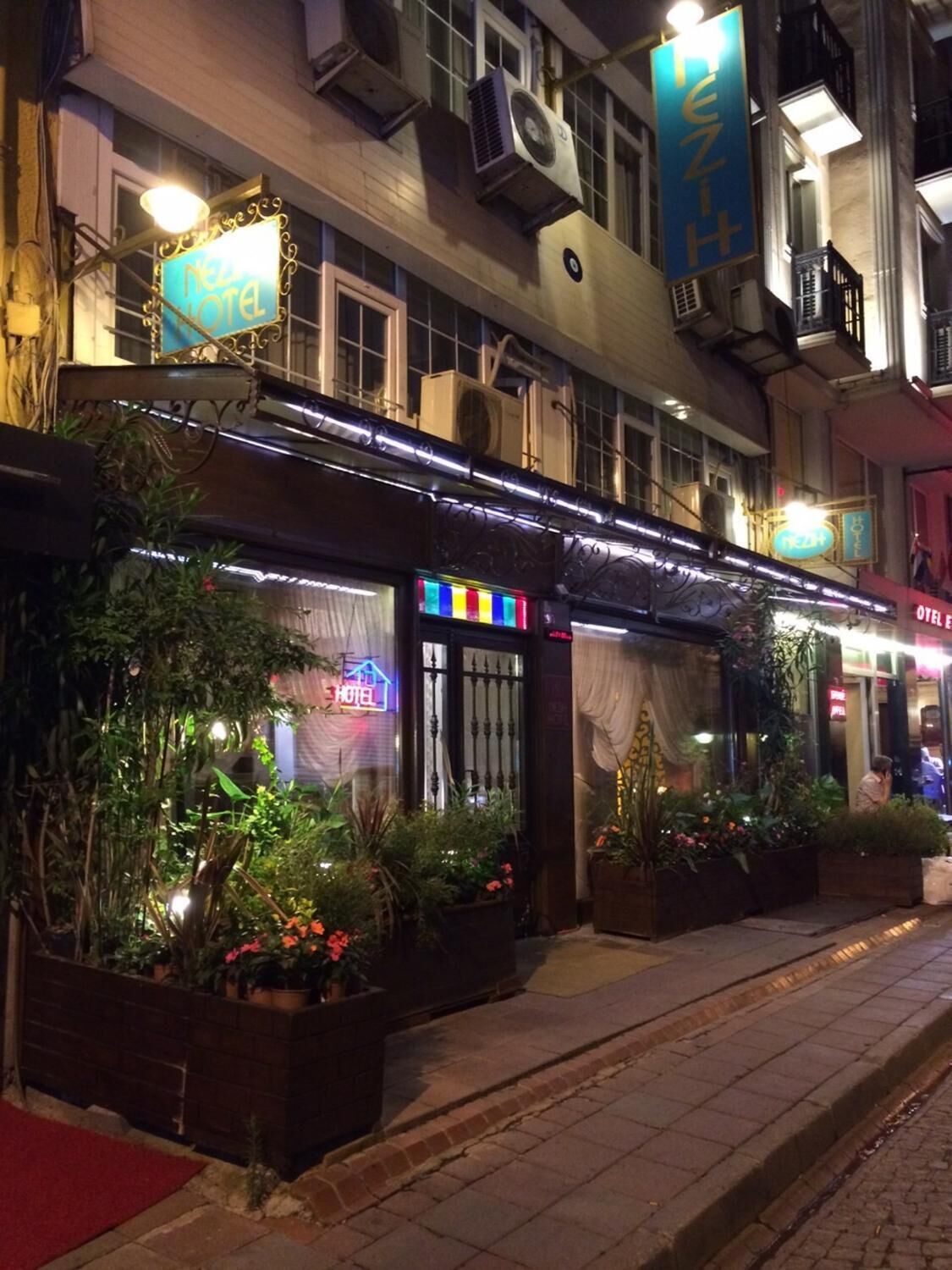 A hotel with a neon sign, outdoor plants, and a red carpet entrance.