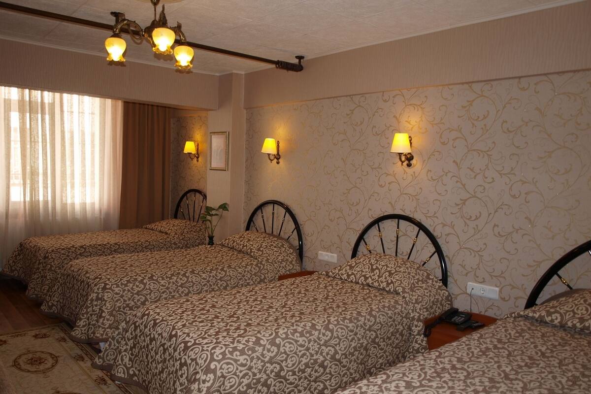 A hotel room with four beds, each with a patterned bedspread. There are wall-mounted lamps, a ceiling light fixture, and a large window with curtains.