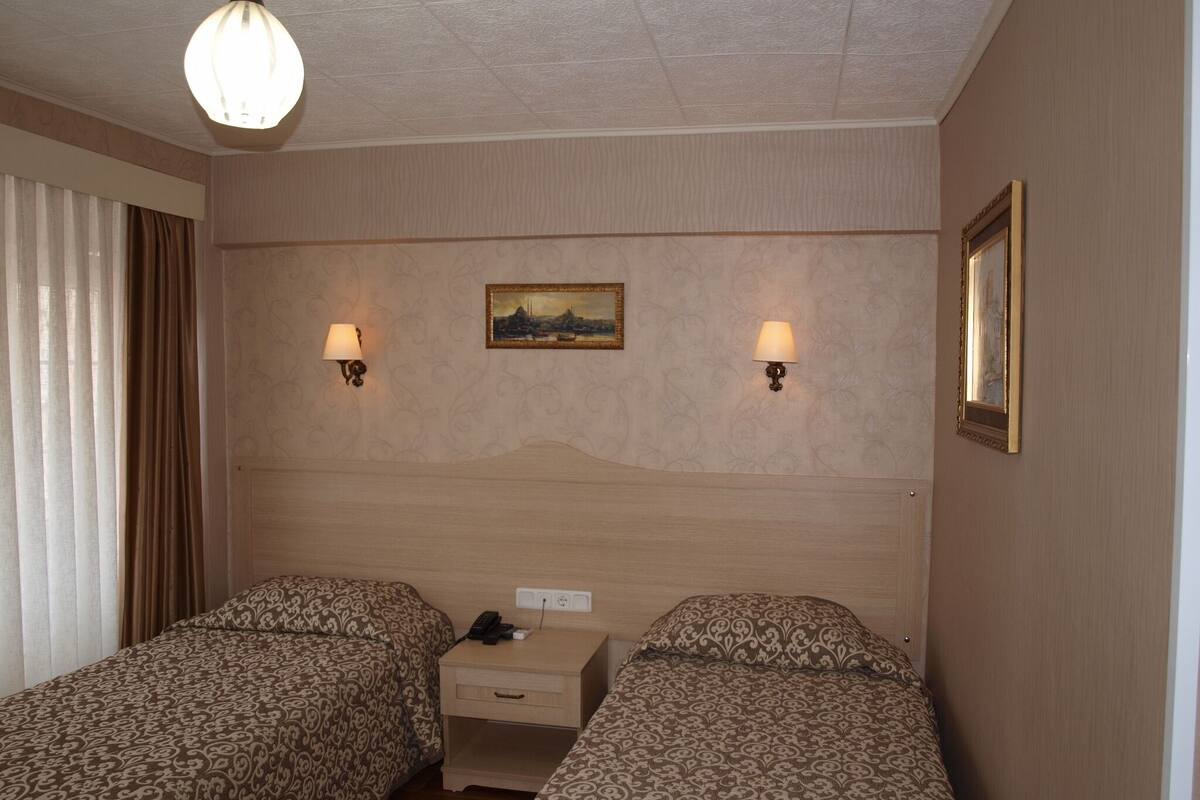 A hotel room with two single beds, a nightstand, a painting on the wall, and two wall-mounted lamps.