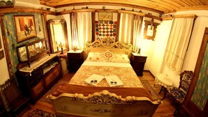 A traditional bedroom with a large bed, ornate headboard, and a quilted wall hanging. There are wooden furniture pieces and a patterned rug on the floor.