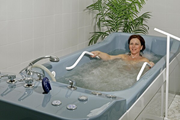 A bathtub with multiple controls and a plant in the background. - Hotel Maxim (Marianske Lazne)