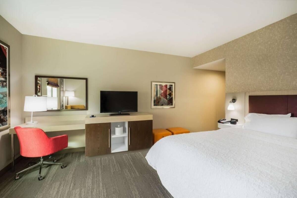 A hotel room with a bed, a desk with a chair, a television, a nightstand with a lamp, and a framed picture on the wall.