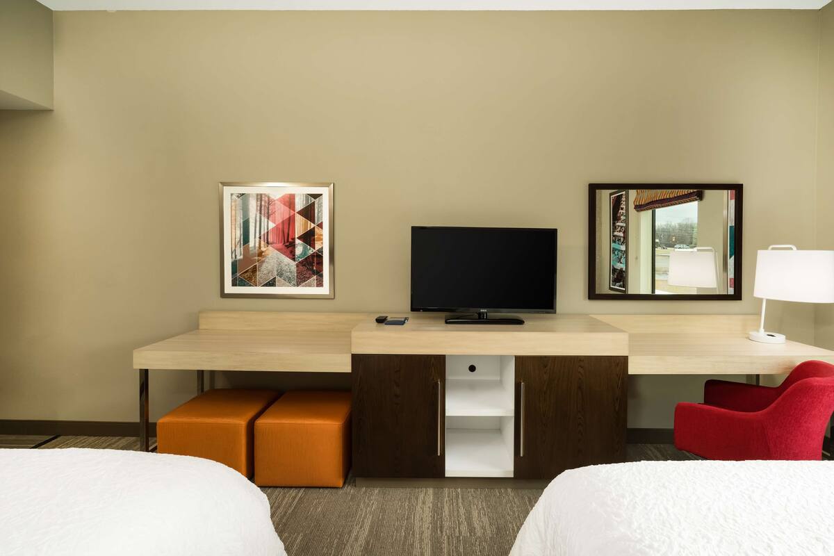 A hotel room with a bed, a desk with a TV, a red chair, a lamp, and artwork on the wall.