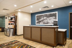 A reception area with a large wall-mounted television displaying a 'smile.' advertisement, a reception desk with a textured panel design, a small kitchenette with a refrigerator and microwave, and a patterned rug on the floor. - Hampton Inn Newport (Newport)