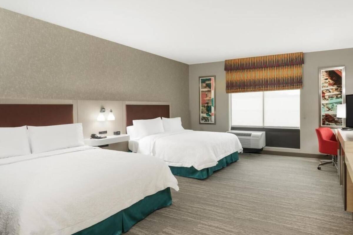 A hotel room with two beds, a desk, a chair, a window with a view, and a painting on the wall.