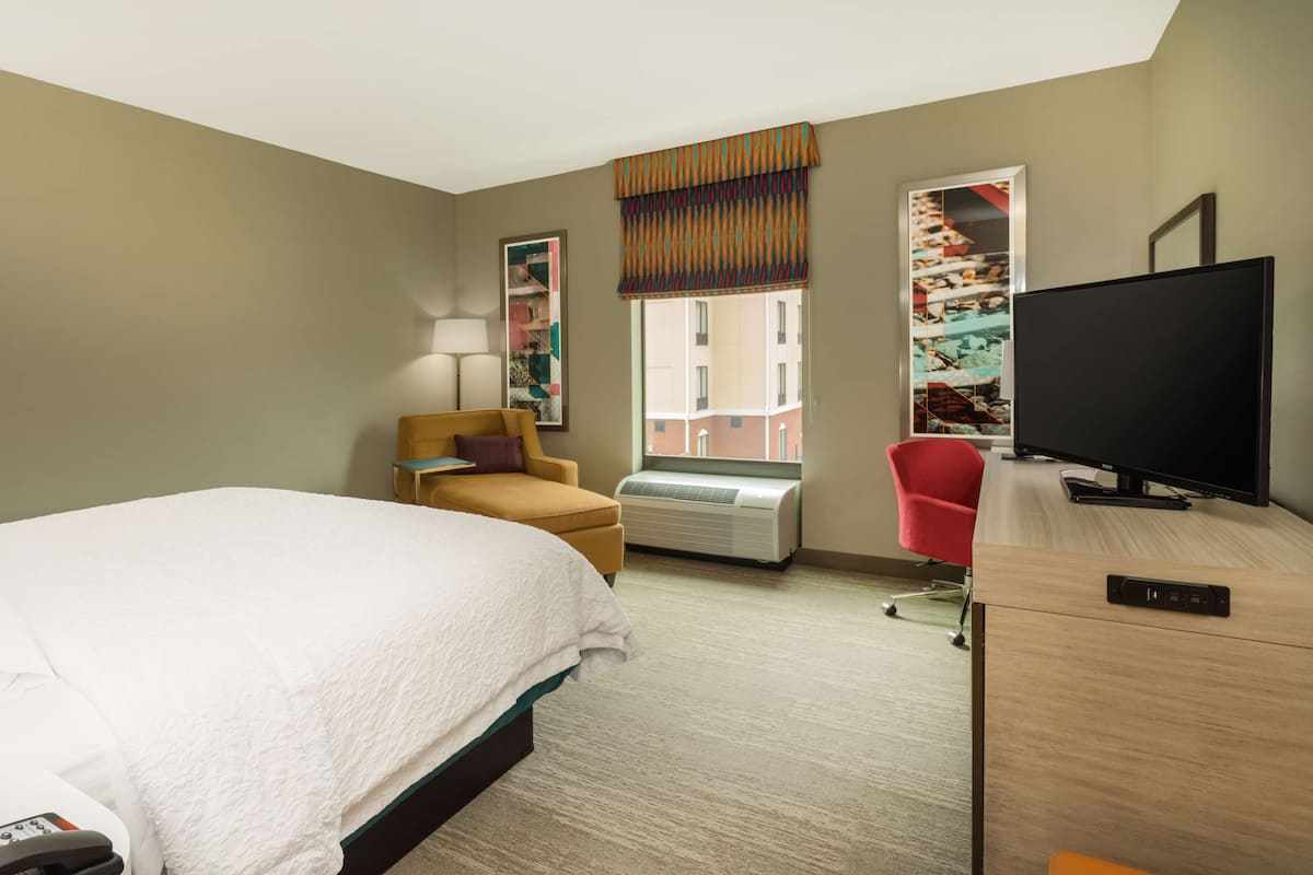 A hotel room with a bed, a desk with a TV, a chair, a lamp, and a window with a view of buildings.