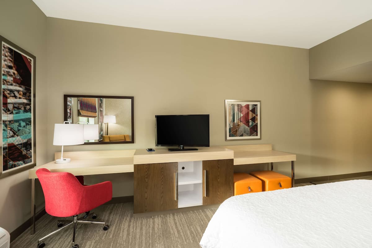 A hotel room with a bed, a desk with a television, a red chair, and two orange storage boxes.