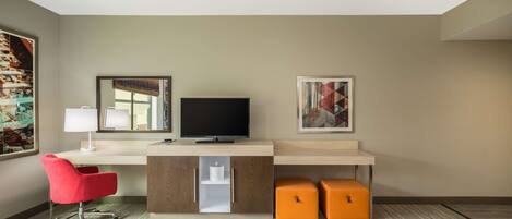 A hotel room with a wooden desk, a red chair, a television, two orange storage boxes, and artwork on the walls.
