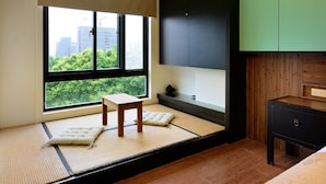 Ein Zimmer mit Tatami-Boden, einem Holztisch, einer Bank und Blick auf eine Stadtlandschaft durch große Fenster.