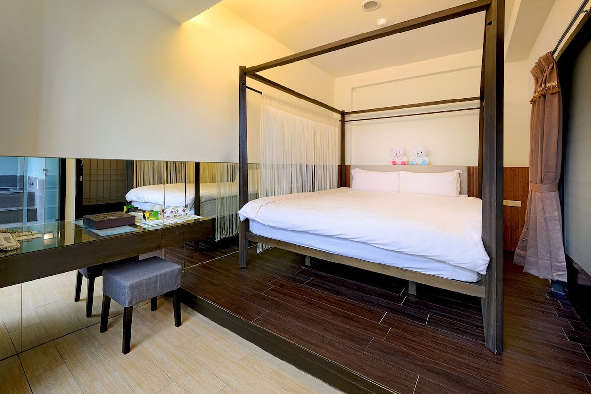 Een moderne hotelkamer met een hemelbed, een glazen bureau met stoel, en een grote spiegel.