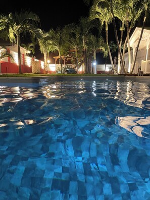 Une piscine nocturne à l'eau claire, dont la surface reflète les lumières, entourée de palmiers et bordée d'un bâtiment aux murs illuminés de rouge et de blanc.