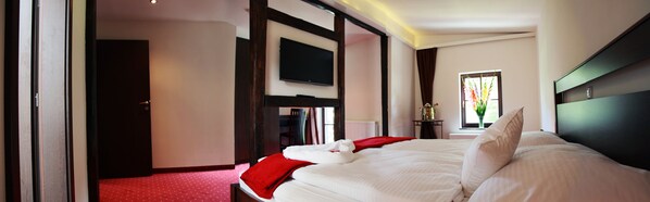 A hotel room with a large bed, a flat-screen TV mounted on the wall, a window with a view of greenery, and a red carpeted floor. - Villa Rozmaryn (Tri Sekery)