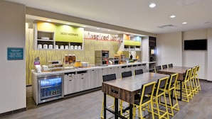 A breakfast area with a self-service station, a large table with chairs, and a refrigerator. - Home2 Suites by Hilton Tampa USF Near Busch Gardens (Tampa)