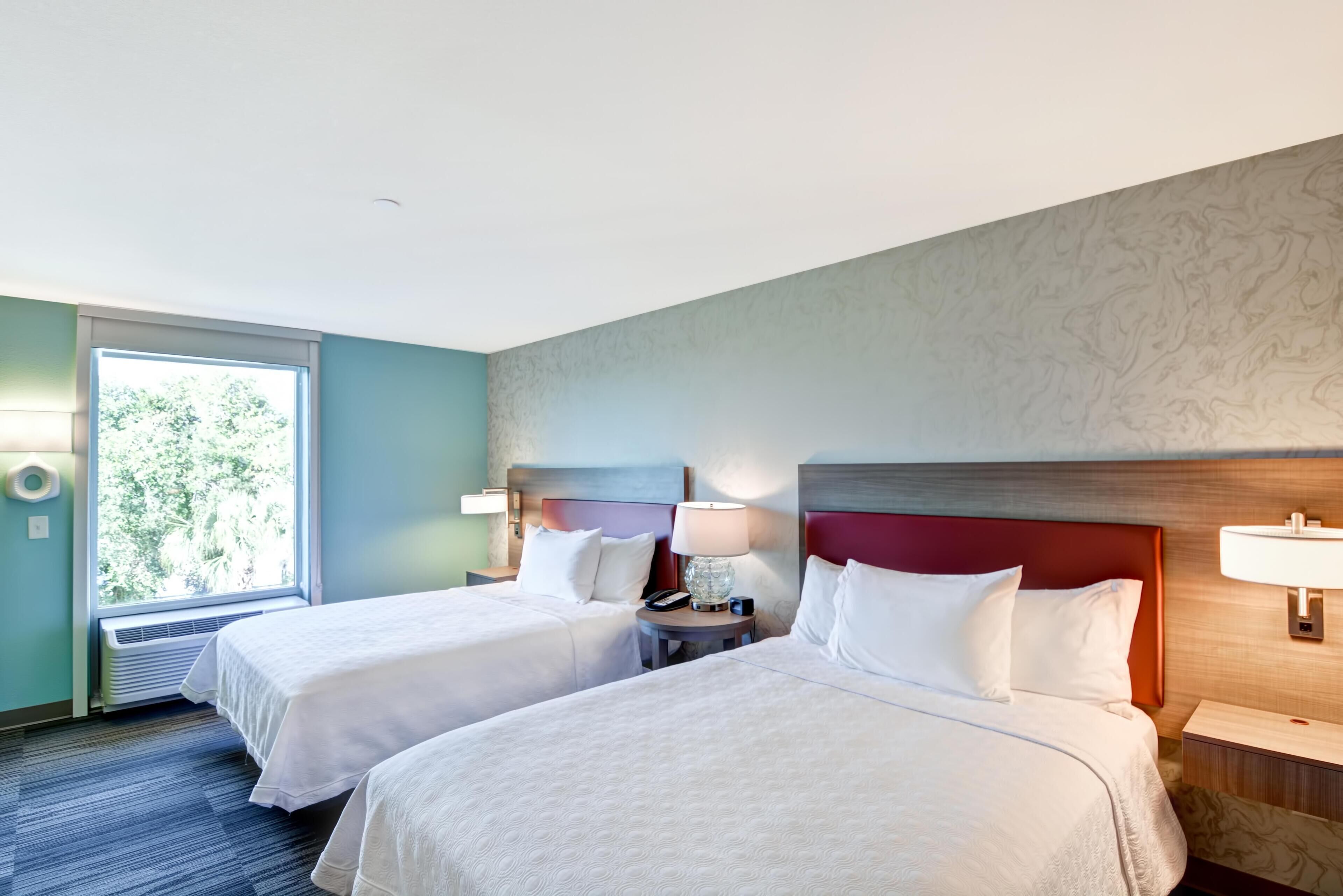 A hotel room with two beds, a window with a view of greenery, a wall-mounted lamp, and a bedside table with a lamp.