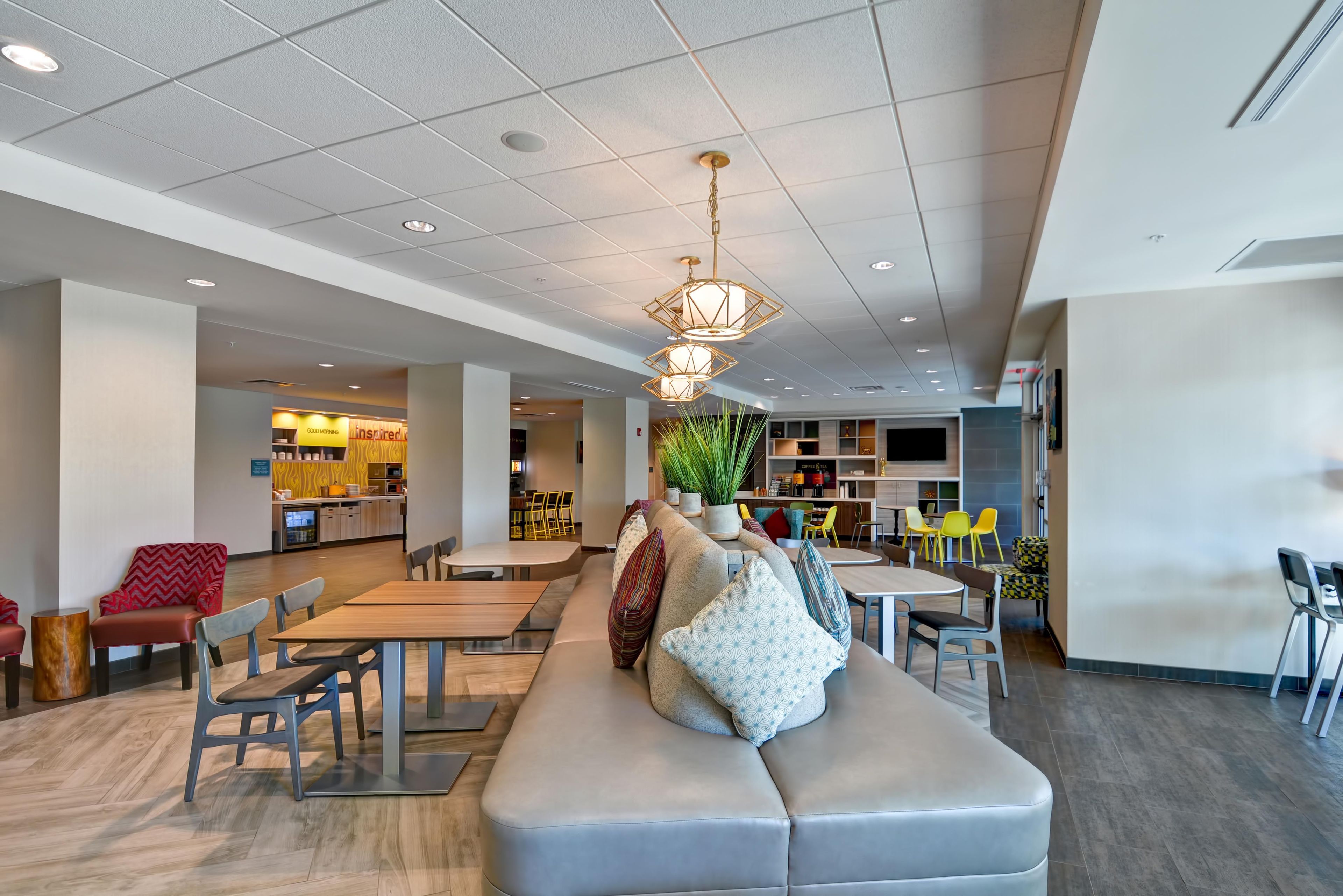 A modern lounge area with a sofa, tables, and chairs, a kitchen area in the background, and a ceiling with recessed lighting and pendant lamps.