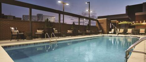 A swimming pool with lounge chairs and umbrellas, surrounded by brick walls and a cityscape view at dusk.