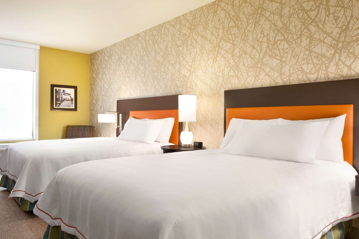 A hotel room with two beds, each with white bedding and orange headboards. There is a framed picture on the wall and a lamp on the nightstand.