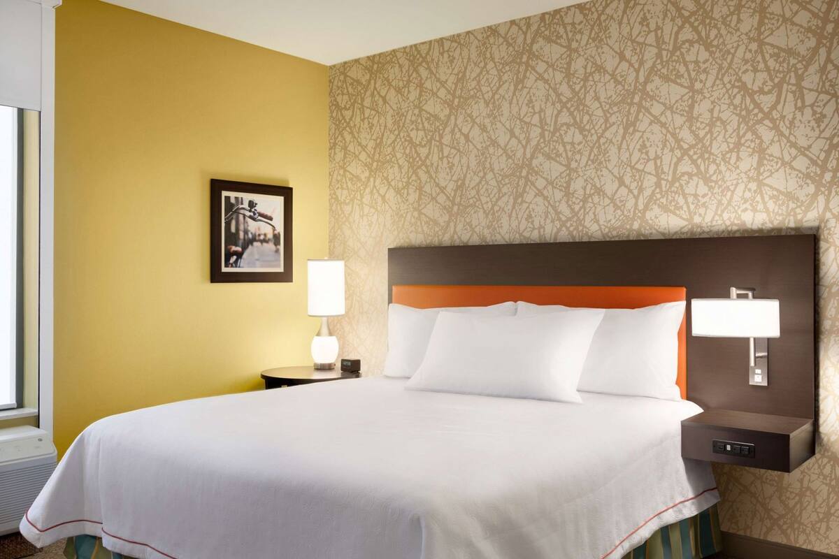 A hotel room with a large bed, a nightstand with a lamp, a framed picture on the wall, and a patterned wallpaper.