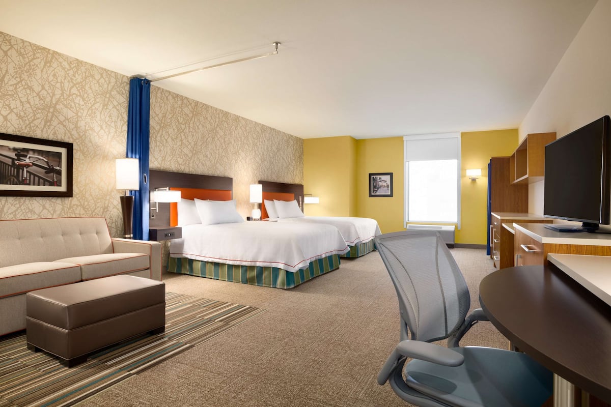 A hotel room with a sofa, two beds, a desk with a TV, and a chair.