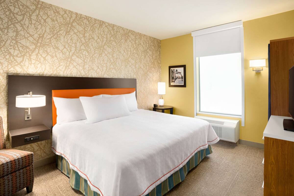 A hotel room with a large bed, a nightstand with a lamp, a small table with a lamp, a framed picture on the wall, and a window with blinds.