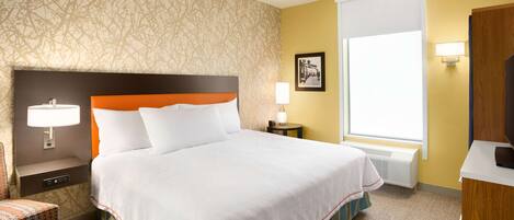 A hotel room with a large bed, a nightstand with a lamp, a small table with a lamp, a framed picture on the wall, and a window with blinds.