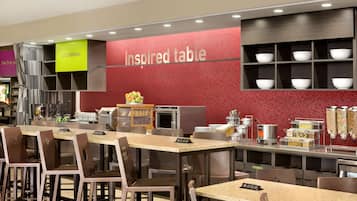 Un restaurant modern amb una paret vermella que diu "Inspired table", una zona de menjar a discreció amb diversos recipients d'aliments i una zona de seients amb taules i cadires.