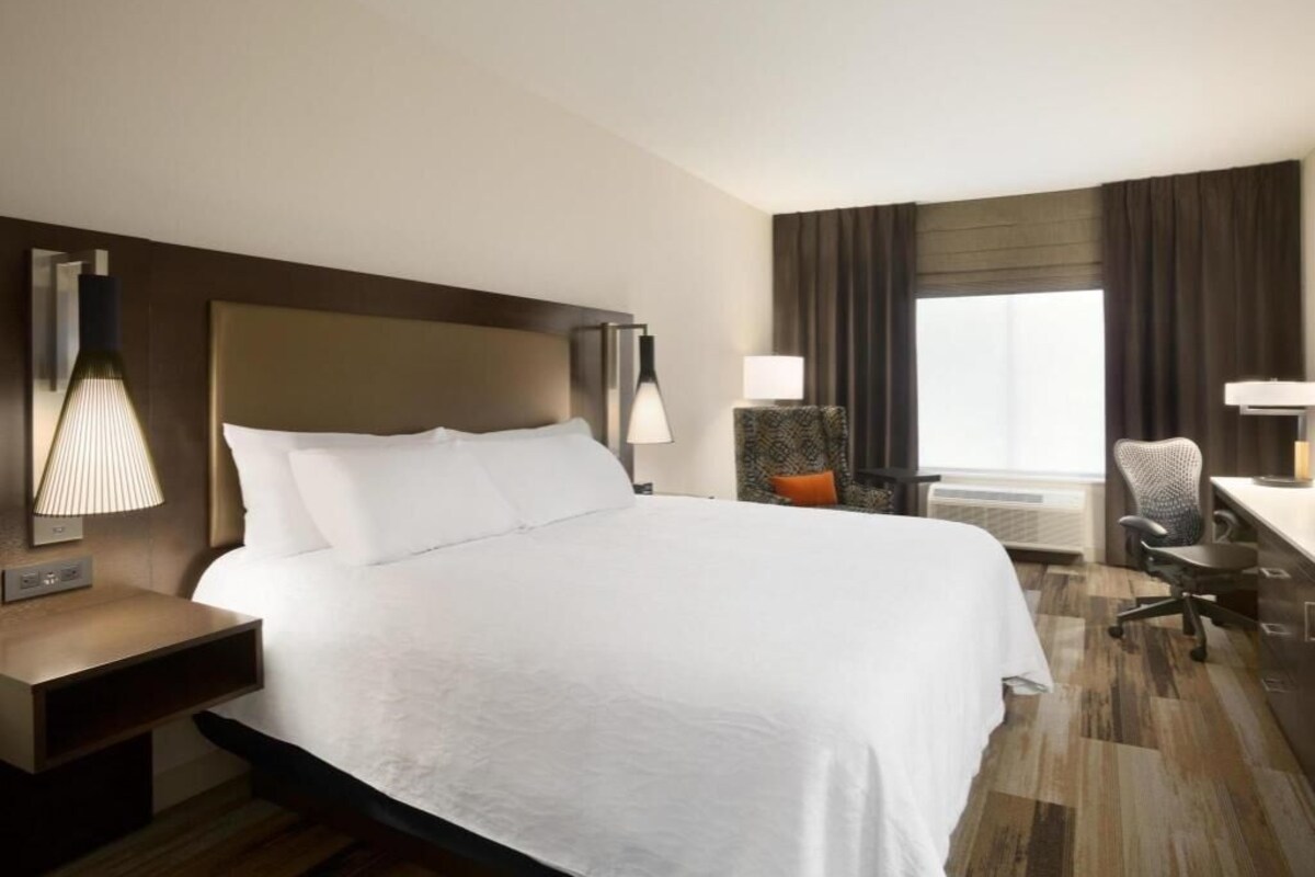 A hotel room with a large bed, bedside lamps, a desk, a chair, and a window with curtains.