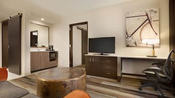 A modern hotel room with a kitchenette, a flat-screen TV, a round coffee table, and a large painting of a bicycle.