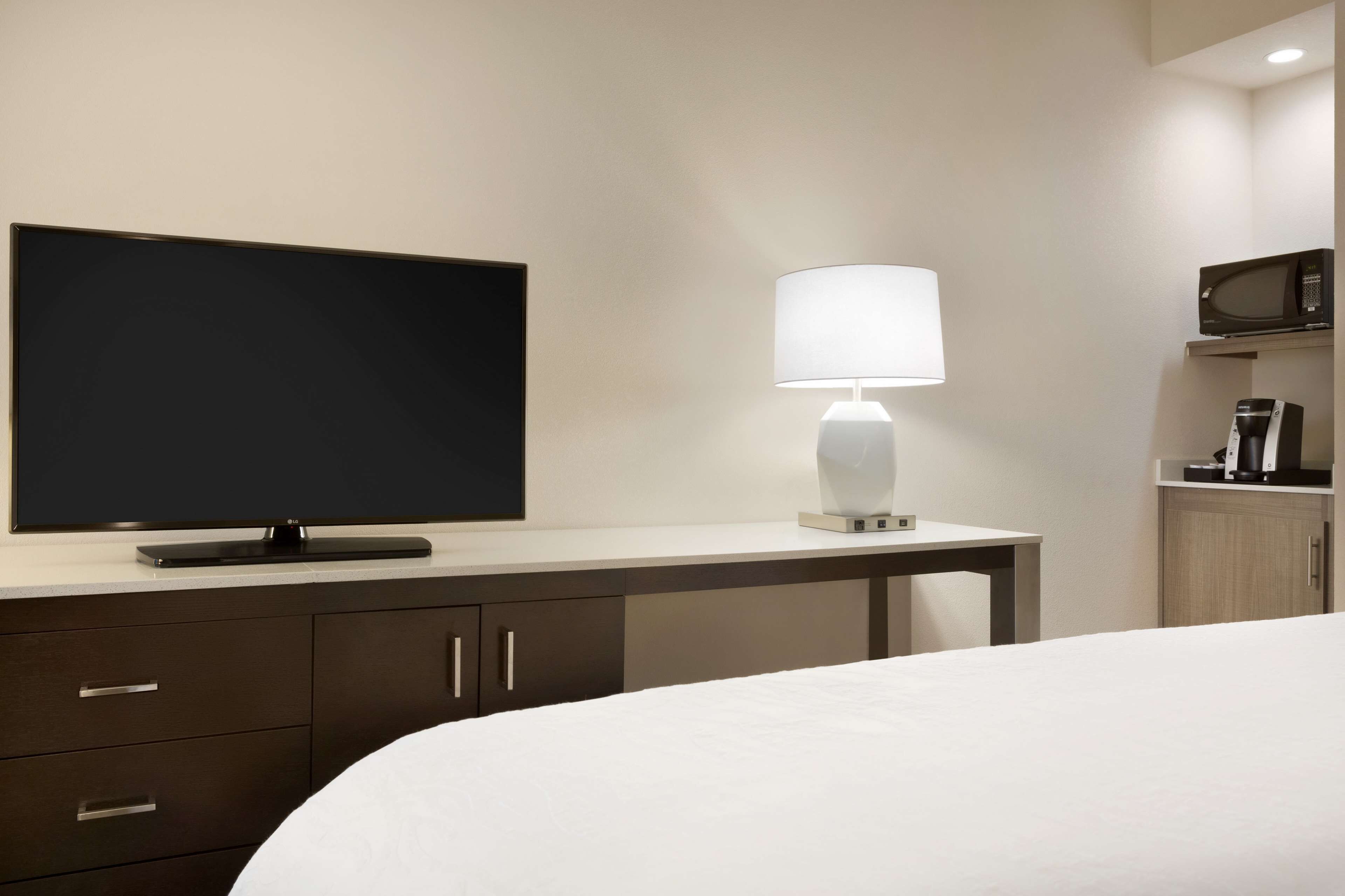 A hotel room with a flat-screen TV on a stand, a bedside table with a lamp, a microwave, and a coffee maker.
