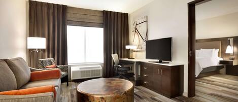 Ein Hotelzimmer mit Sofa, Sesseln, rundem Couchtisch, Schreibtisch, Fernseher und einem Bett mit Kopfteil.