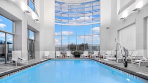 Piscina interna com paredes de vidro transparente, cadeiras de lounge brancas e vista para o céu.