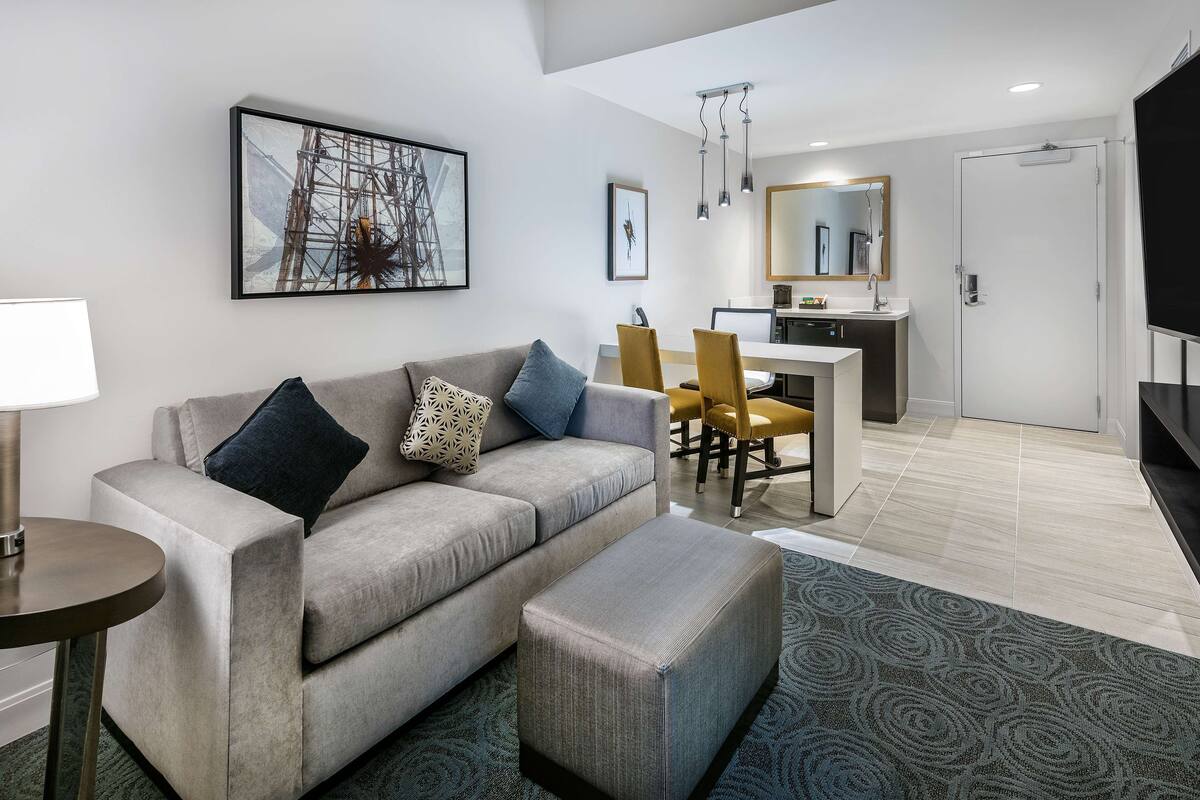 Una habitación de hotel moderna con sofá, reposapiés y un área de comedor con sillas y una mesa.
