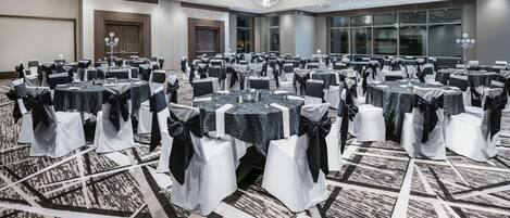 A large conference room with multiple round tables, each with a white tablecloth and black chair covers. The room has a carpeted floor with a geometric pattern and large windows along one wall.
