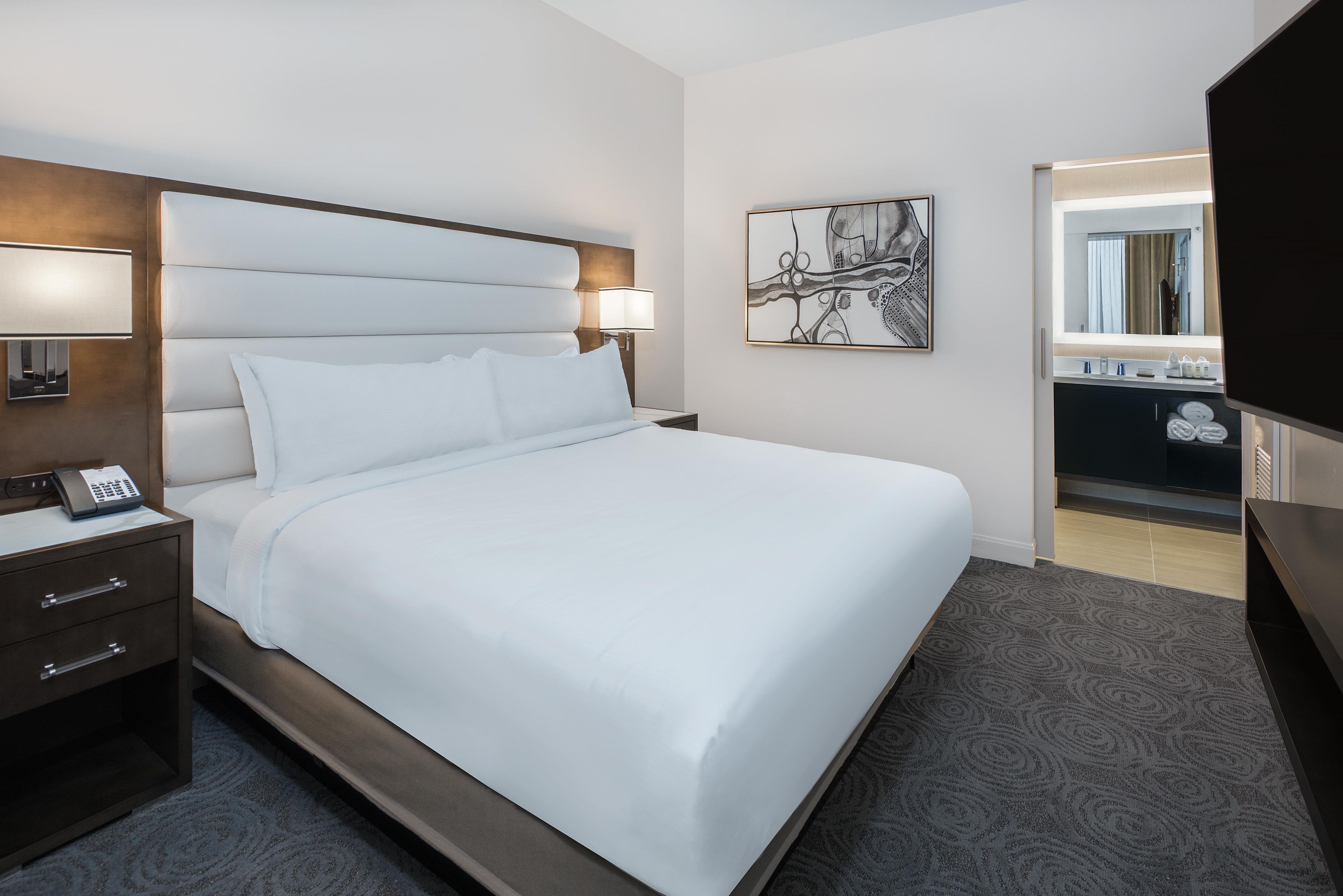 A hotel room with a large bed, a bedside table with a phone, a wall-mounted lamp, and a painting.