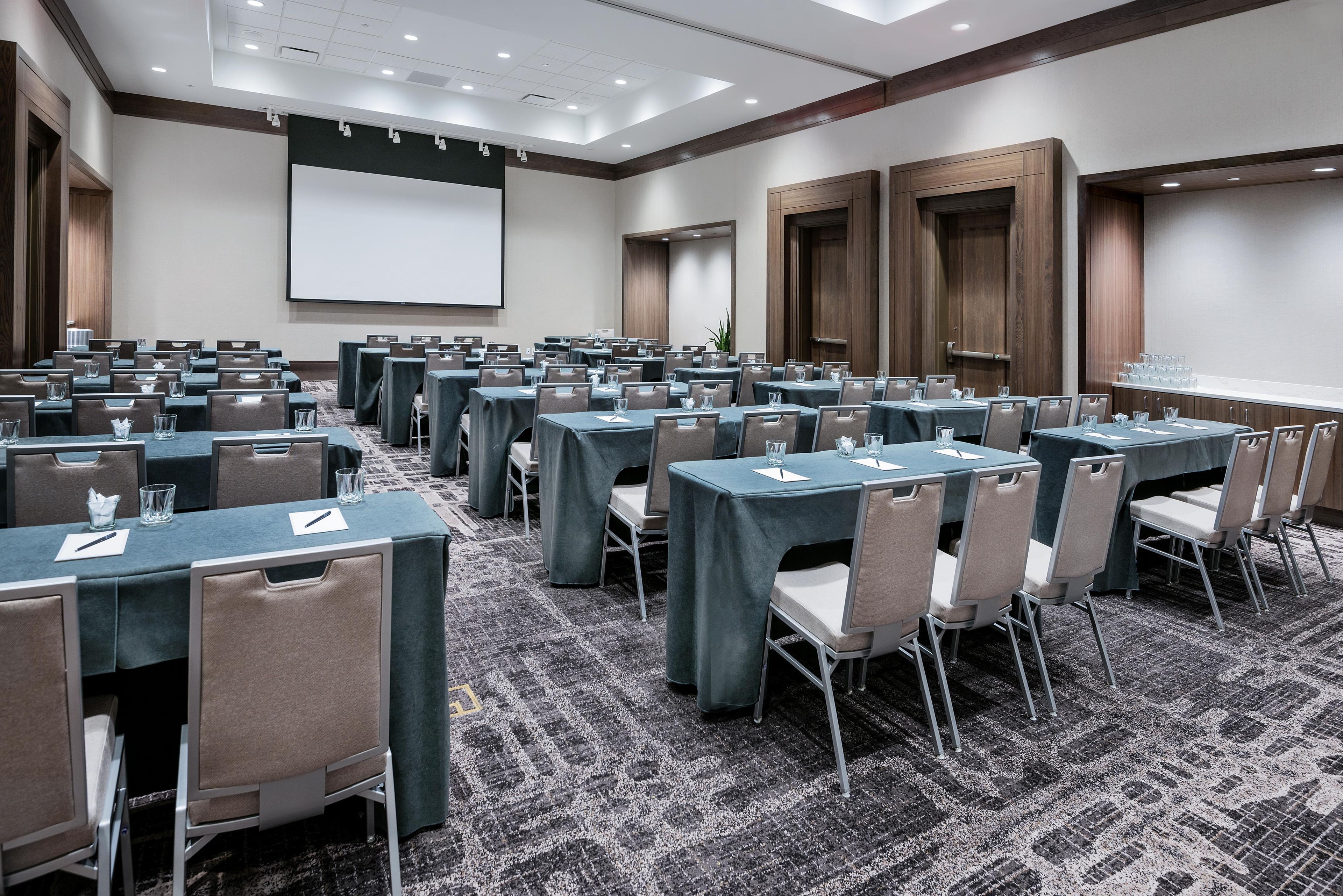 A conference room with multiple tables and chairs, a projector screen, and a carpeted floor.