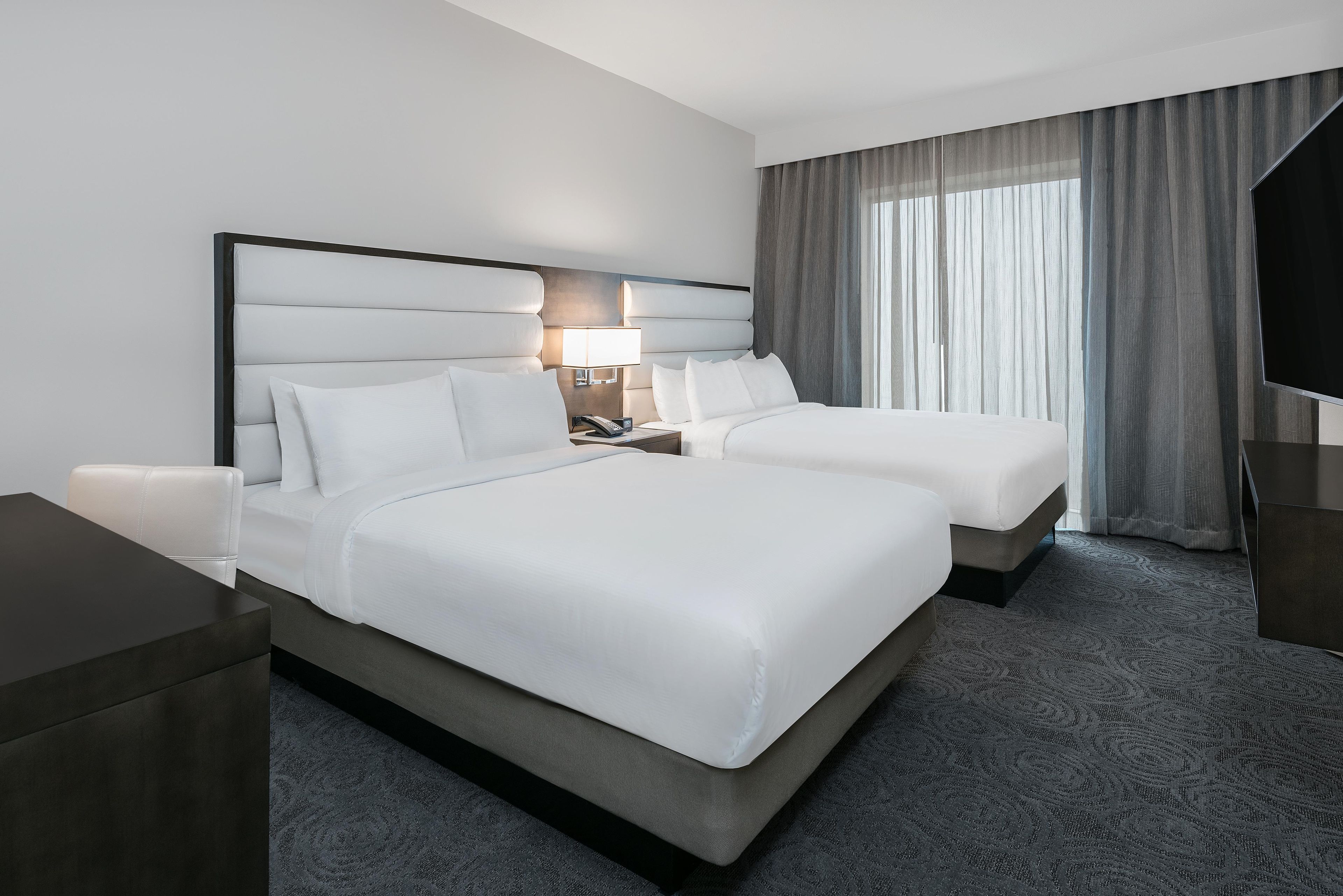 A hotel room with two beds, a bedside table, a lamp, and a large window with curtains.