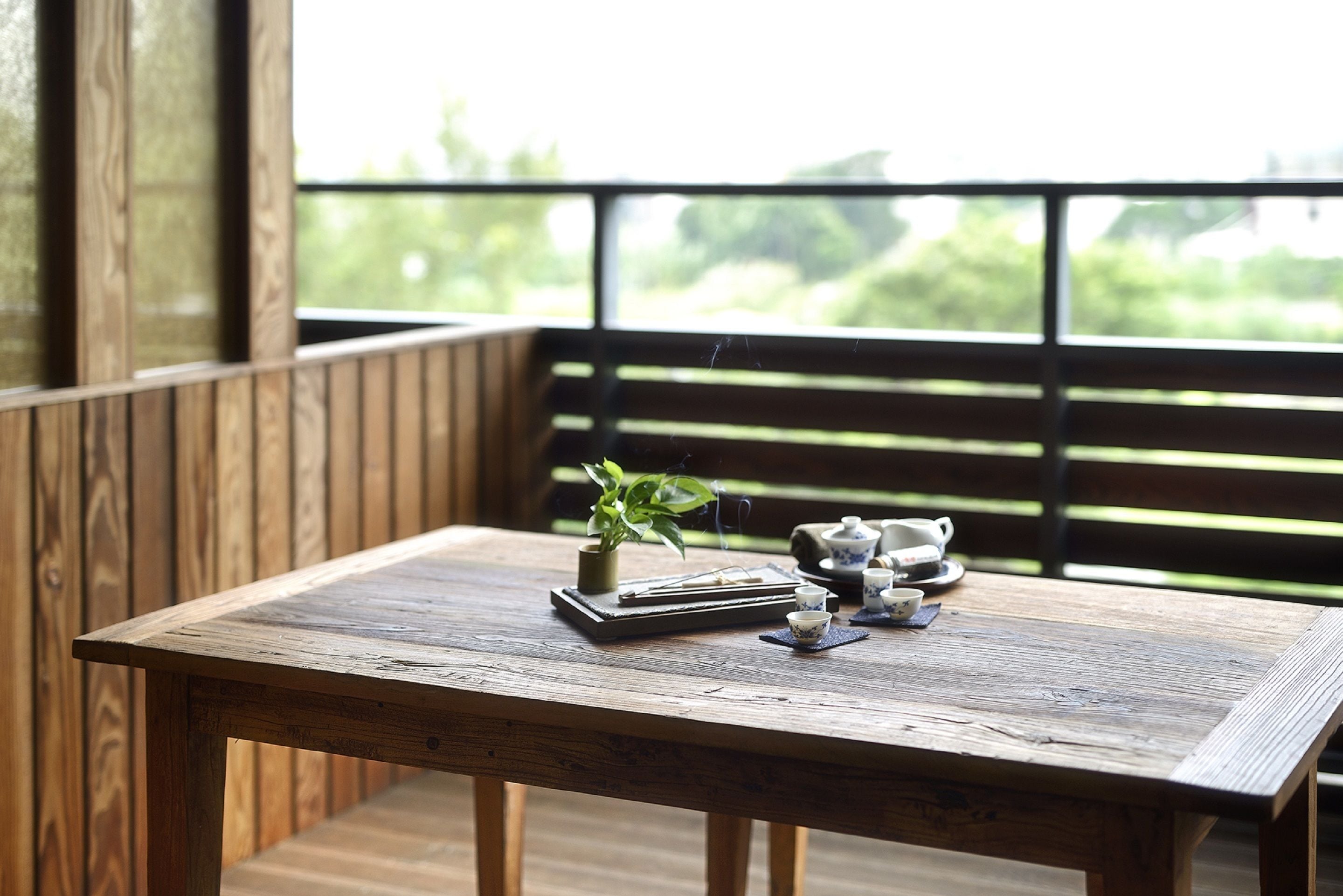 A wooden table with a plant, teapot, cups, and a tray on a balcony with wooden railings and a view of greenery.